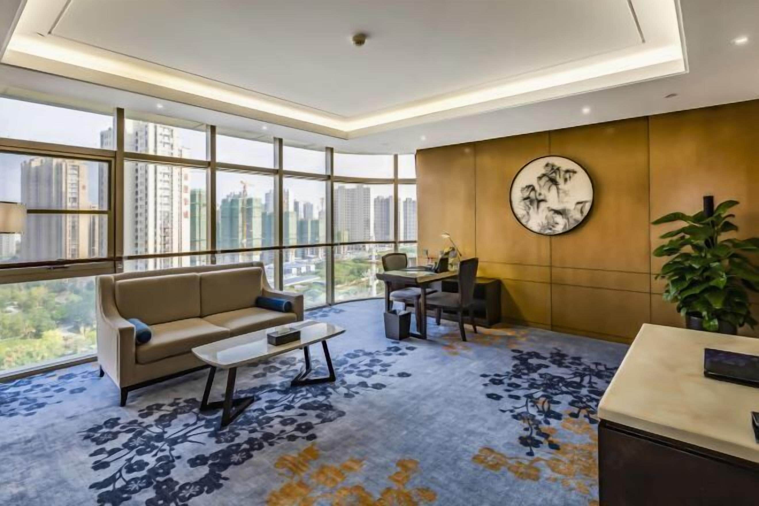A modern office space with a sofa, desk, and chair, large windows with a city view, and a round wall decoration.