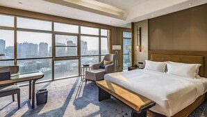 A modern hotel room with a large bed, a desk, a chair, and a view of the cityscape through large windows. - Shangqiu Xinhua Jianguo Hotel (Shangqiu)