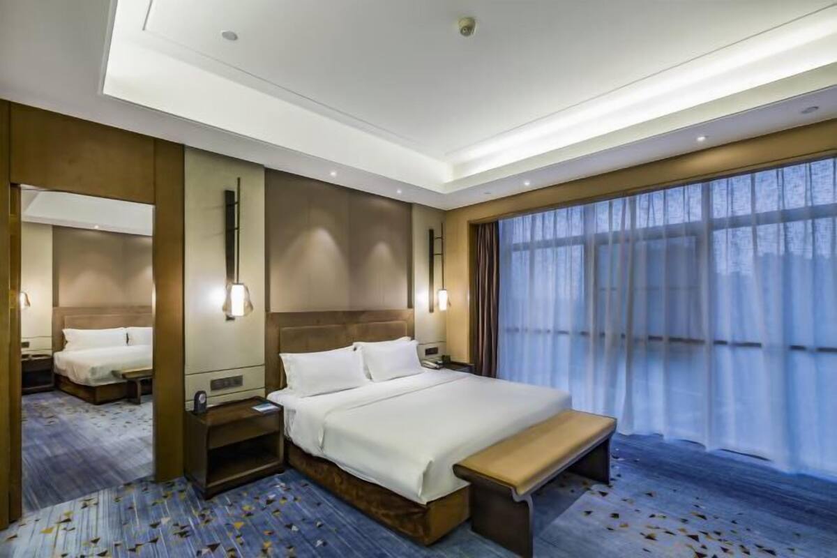 A modern hotel room with a large bed, bedside tables, a bench, and a large window with curtains.