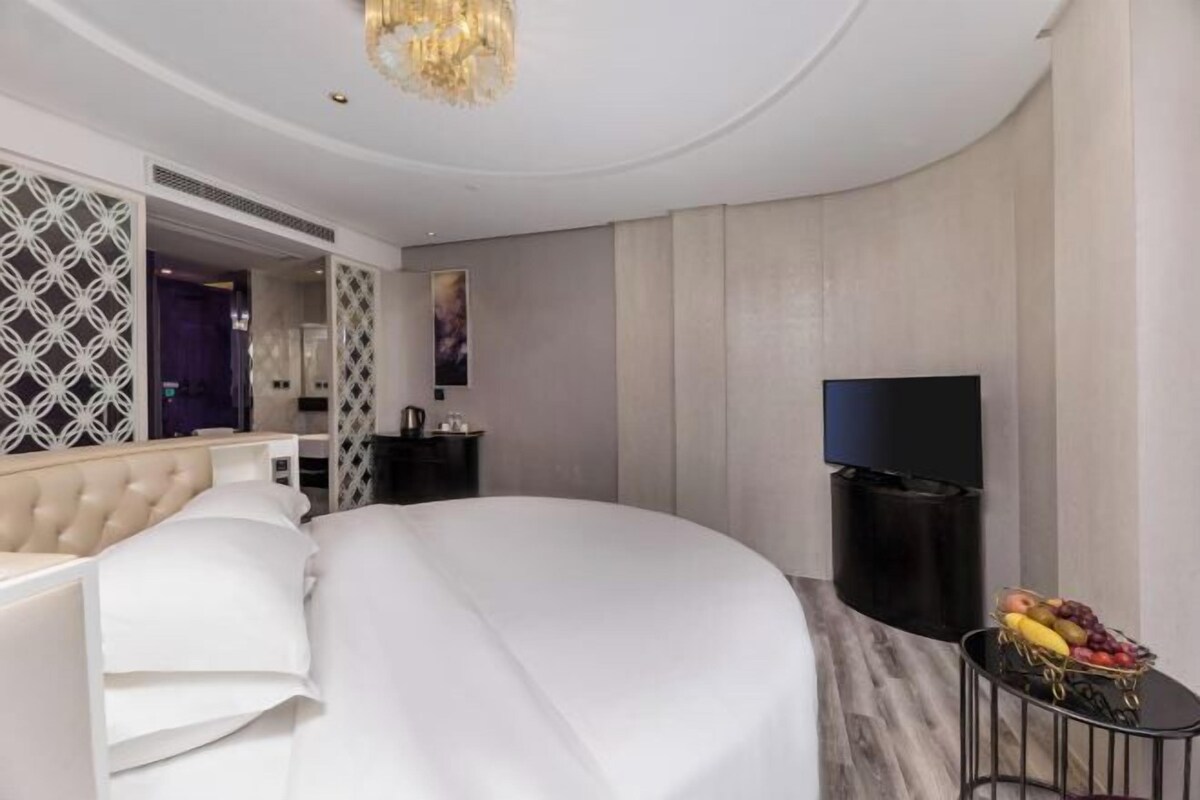 Ein modernes Hotelzimmer mit einem großen Bett, einem Fernseher, einer Obstkorb und einem dekorativen Spiegel.
