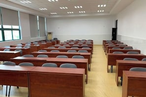 Une salle de classe avec des rangées de bureaux et de chaises, un écran de projection et une grande fenêtre.