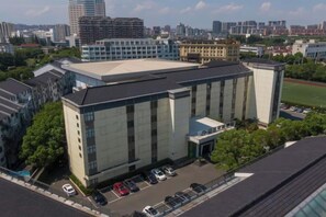 A modern building with a large glass facade, a parking lot, and a sports field in the background. - China Youth Hostel Landscape Garden Hotel (Suzhou Wujiang RT-Mart) (Suzhou)