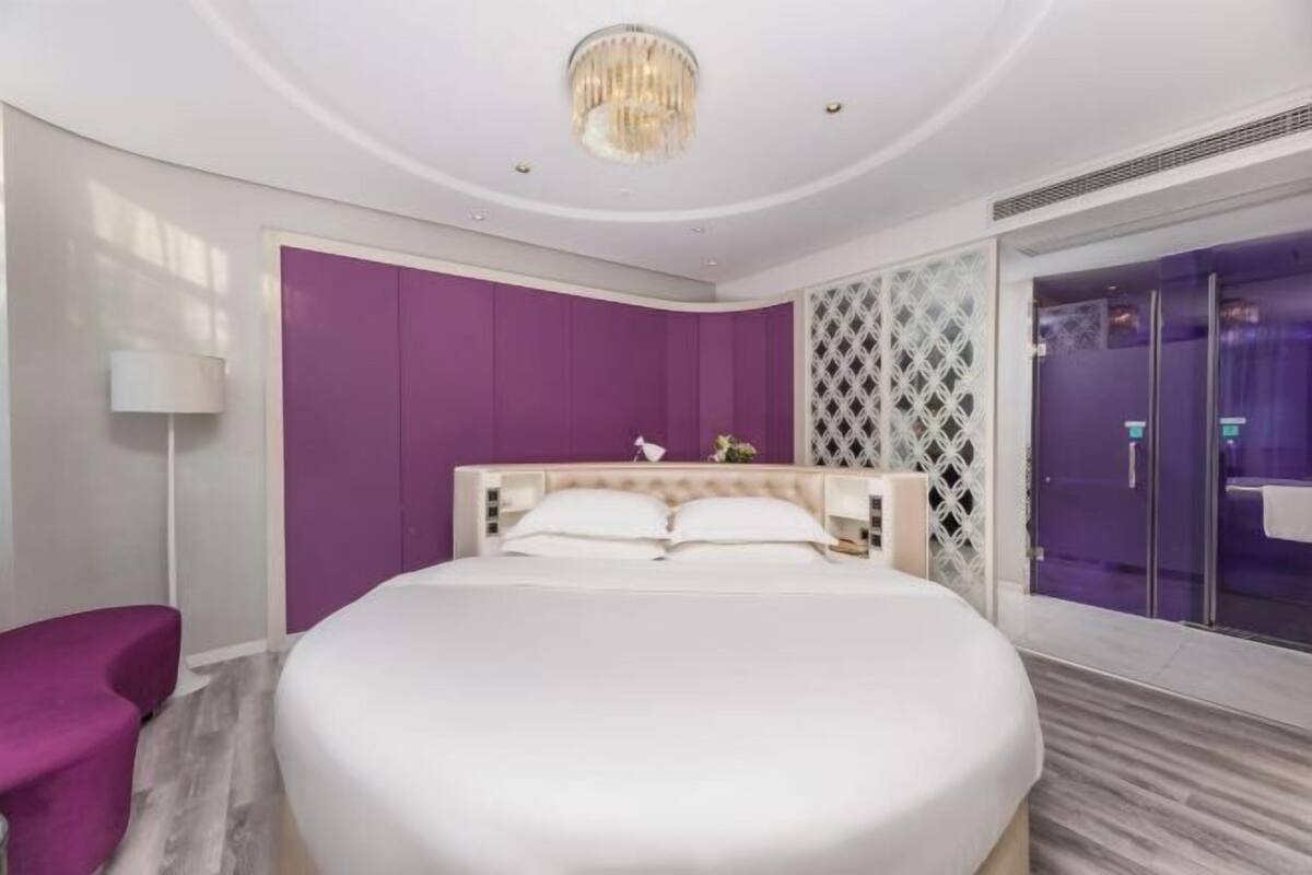 Un dormitorio moderno con una cama grande, una pared de acento morada, una araña y una zona de ducha de vidrio.