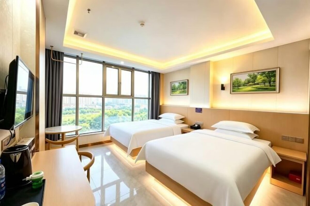 A hotel room with two beds, a desk, a chair, a TV, and a large window offering a city view.