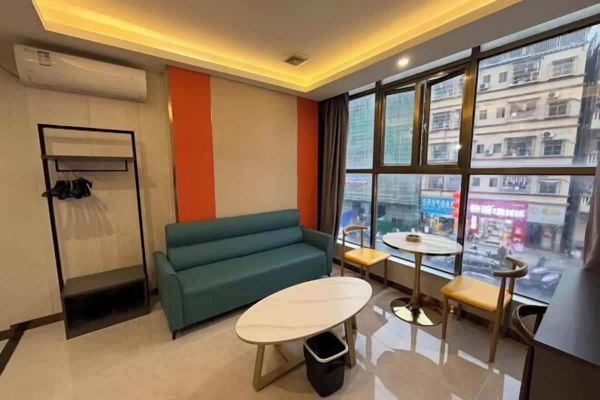 A modern hotel room with a sofa, a small table, a chair, a window with a view of buildings, and a wall-mounted air conditioning unit.