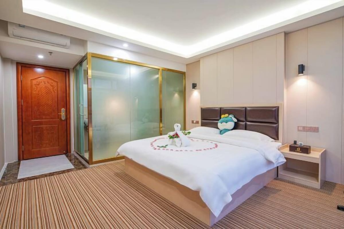 A hotel room with a large bed, a wooden door, a glass partition, and a nightstand with a lamp.