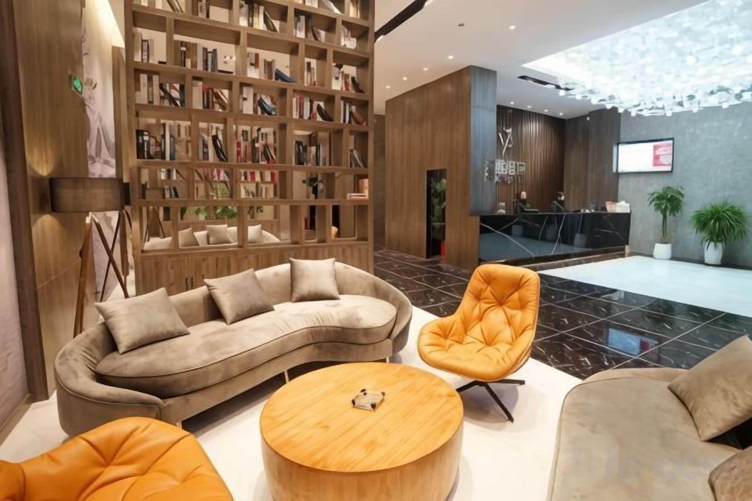 A modern lobby with a curved sofa, a round wooden coffee table, and a seating area with an orange chair. A large bookshelf filled with books is visible, and a reception desk is in the background.