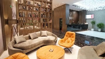 A modern lobby with a curved sofa, a round wooden coffee table, and a seating area with an orange chair. A large bookshelf filled with books is visible, and a reception desk is in the background.