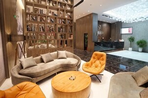 Un vestíbulo moderno con un sofá curvo, una mesa de centro redonda de madera y una zona de estar con una silla naranja. Se ve una estantería grande llena de libros, y un mostrador de recepción al fondo.