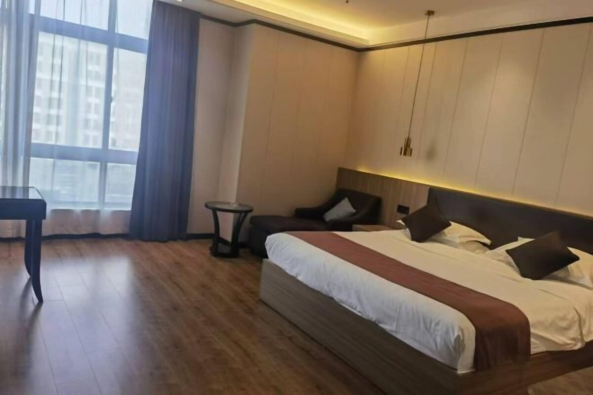 A hotel room with a large bed, a small round table, a chair, and a view of buildings through a window.