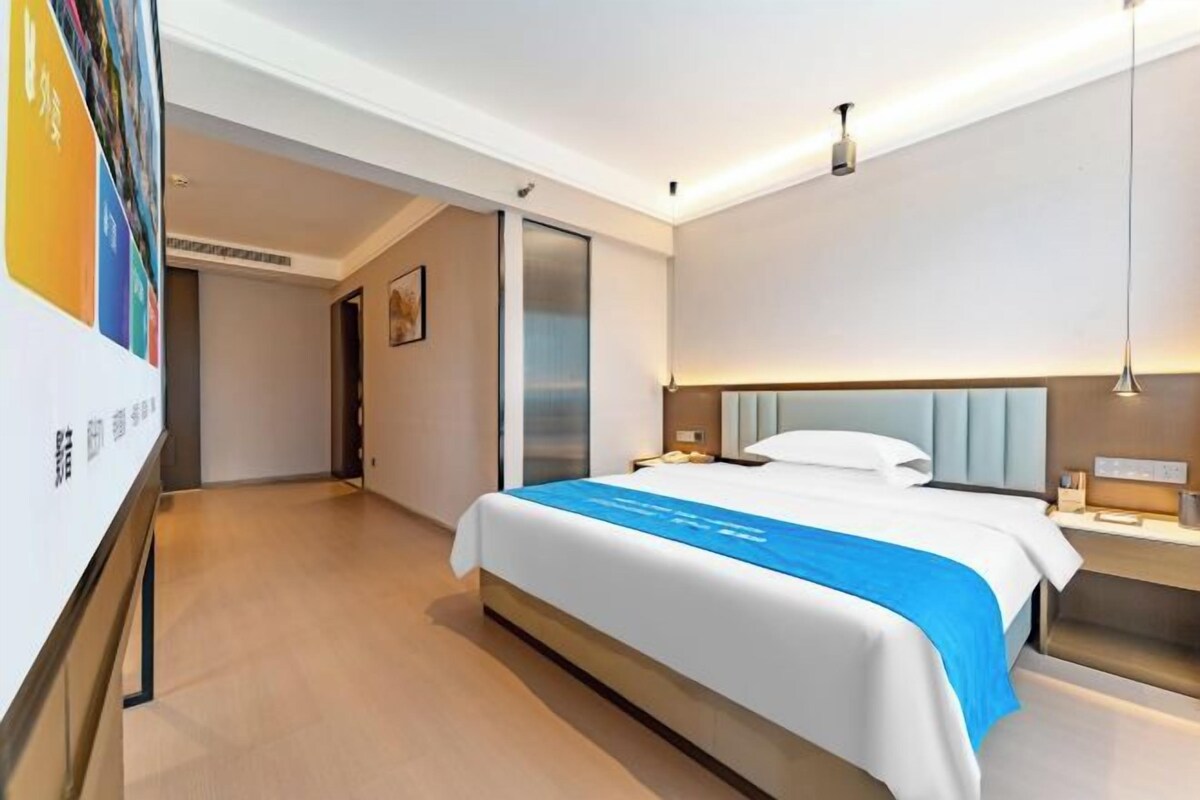 A modern hotel room with a large bed, bedside tables, a nightstand, and a wall-mounted lamp.