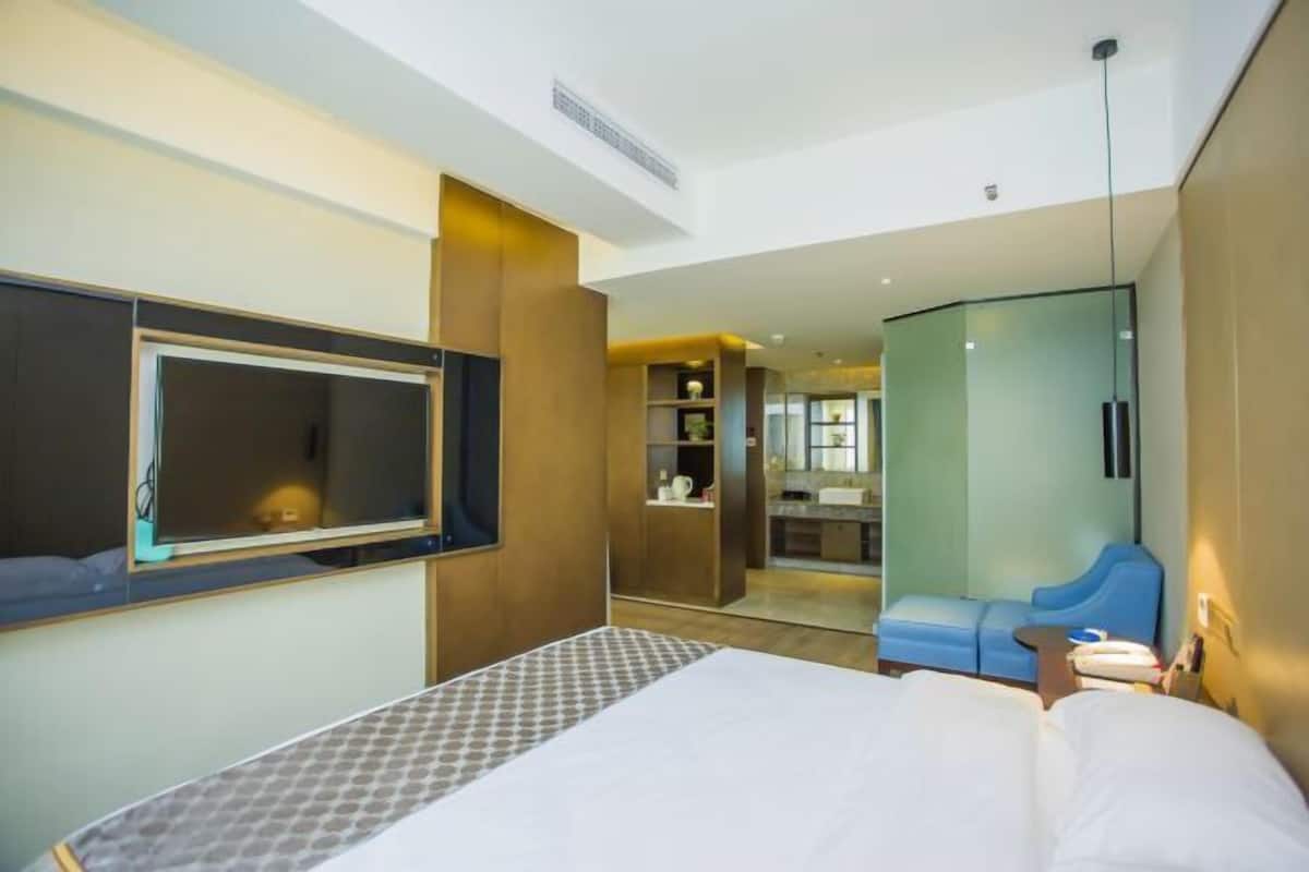 A modern hotel room with a large bed, a TV, a seating area, and a glass-enclosed bathroom.