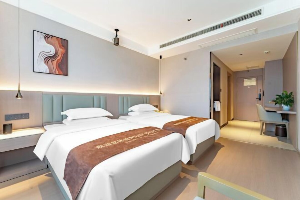 A hotel room with two beds, a desk, a chair, and a small plant.