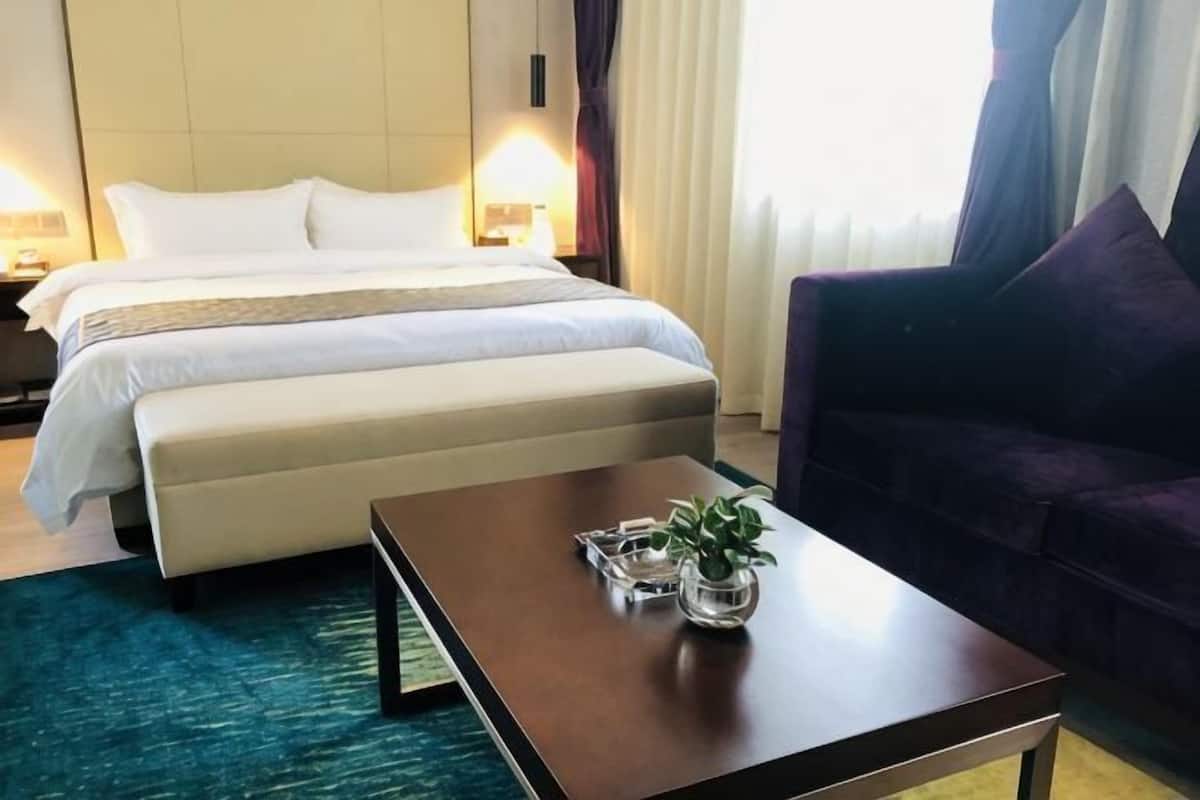 A hotel room with a bed, a sofa, a coffee table with a plant, and a window with curtains.