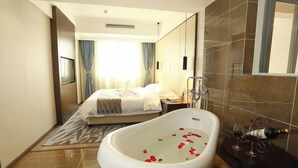 A hotel room with a large bathtub filled with rose petals, a bed with a white comforter, a bedside table with a bottle and glasses, and a large mirror on the wall. - Xinshenghao Hotel (Chengdu)