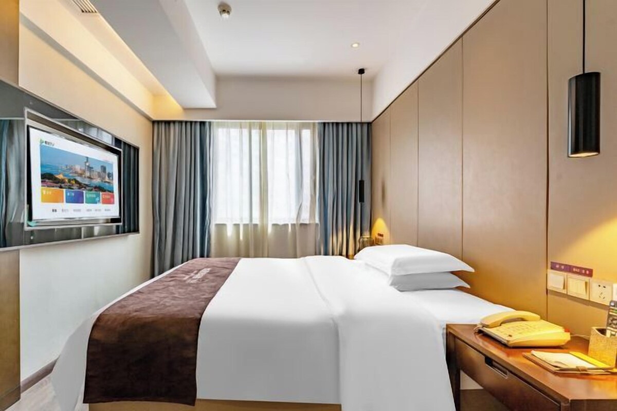 A hotel room with a bed, a television mounted on the wall, a desk with a telephone, and a window with curtains.