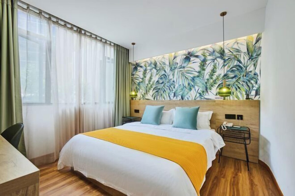 A hotel room with a large bed, a wooden headboard, a tropical leaf mural, a desk, and a chair.