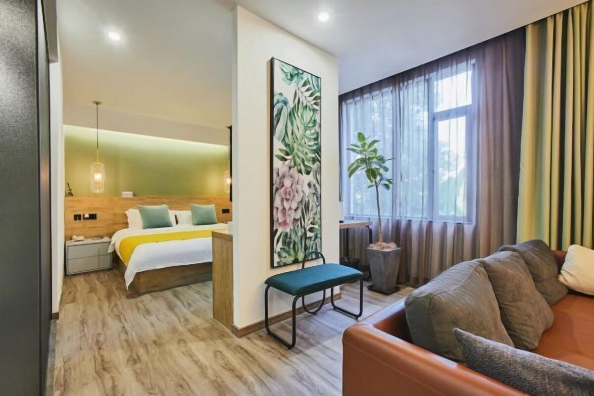 A modern hotel room with a bed, a green bench, a sofa, and a large plant.