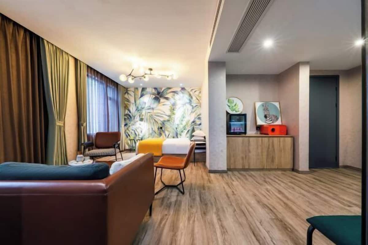 Habitación de hotel con cama, sofá, sillón, una mesita, televisor y un mural tropical en la pared.