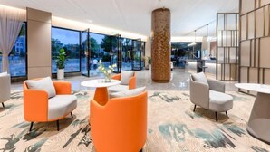 Eine moderne Lobby mit orangefarbenen und grauen Sesseln, einer großen Säule und bodentiefen Fenstern mit Blick ins Freie.