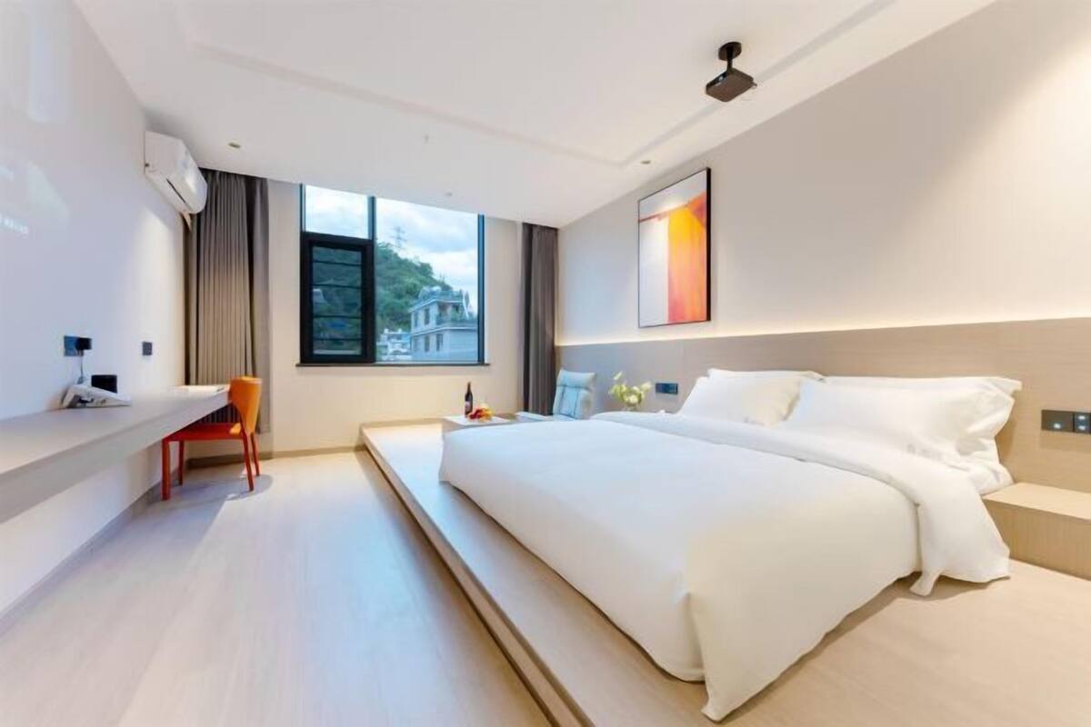 Ein modernes Hotelzimmer mit einem großen Bett, einem Schreibtisch und einem Sessel. Ein Fenster bietet Blick auf die Stadt.