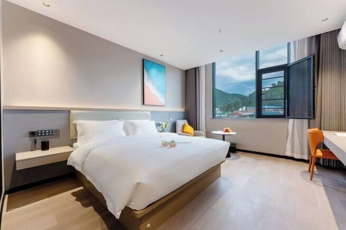 Ein modernes Hotelzimmer mit einem großen Bett, einem Nachttisch und Blick auf die Stadt durch ein großes Fenster.