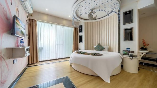 A modern hotel room with a curved bed, a large window with curtains, a ceiling with a decorative design, and a small desk with a chair.