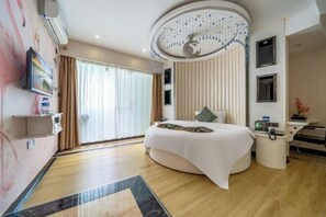 A modern hotel room with a curved bed, a large window with curtains, a ceiling with a decorative design, and a small desk with a chair.