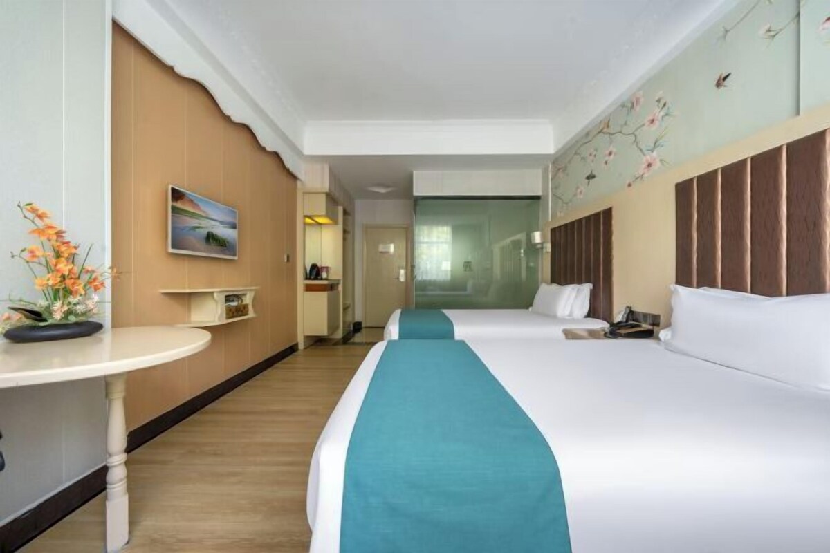 A hotel room with two beds, a small table with a vase of flowers, a painting on the wall, and a view through a glass door.