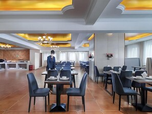 Comedor de hotel con mesas y sillas, un camarero y una cocina al fondo.