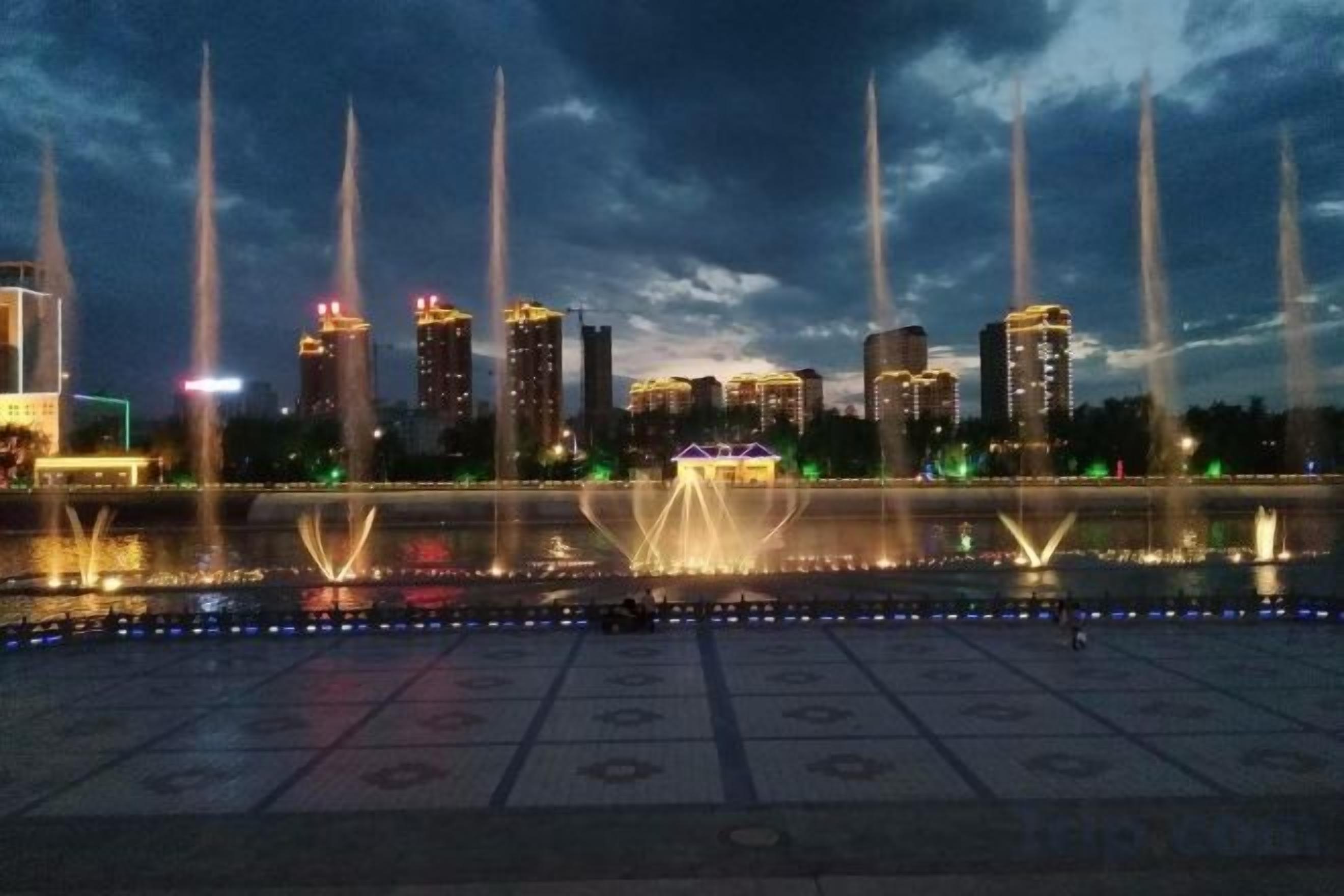 Ein beleuchtetes Stadtbild bei Nacht mit Gebäuden und Springbrunnen.