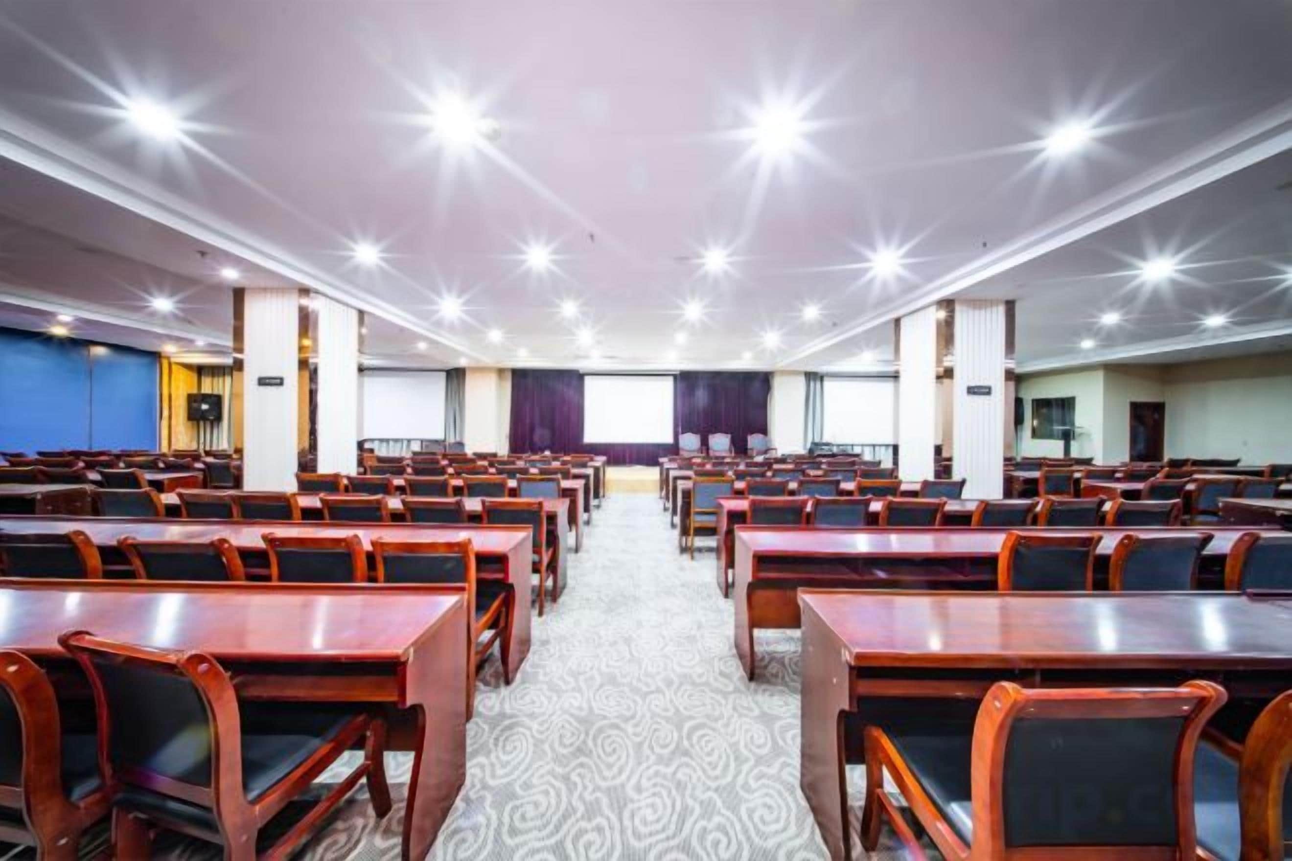 Un salón de conferencias con mesas y sillas dispuestas en filas, piso alfombrado y un amplio ventanal con cortinas.