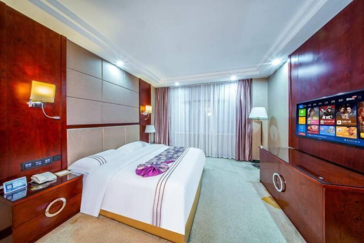 A hotel room with a large bed, a TV mounted on the wall, a desk with a lamp, and a wooden wardrobe.