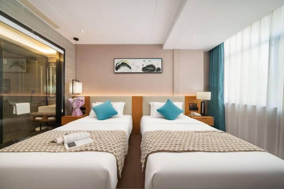 A hotel room with two beds, a bedside table with a lamp, a wall-mounted artwork, and a window with curtains.