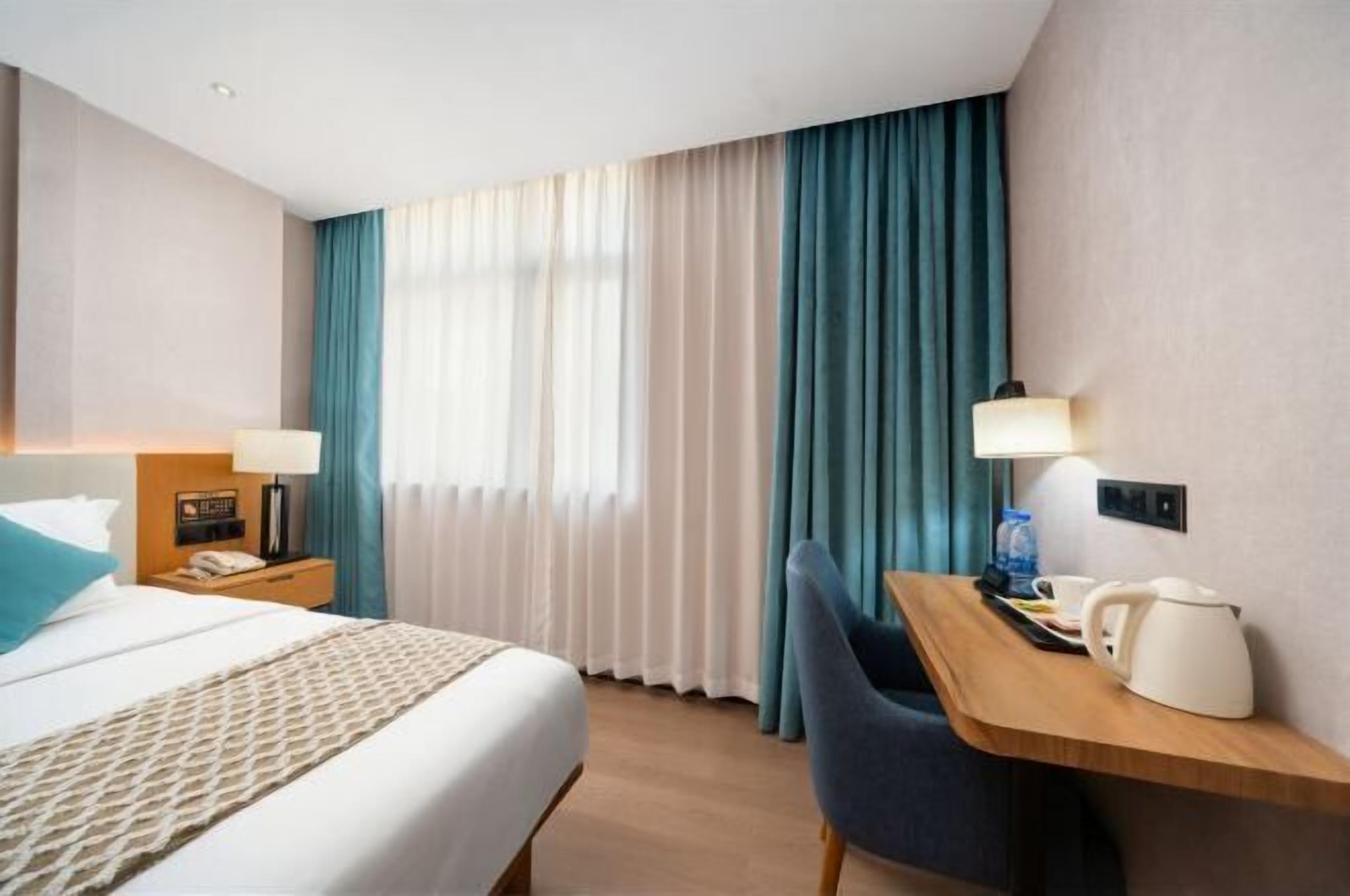 Une chambre d’hôtel avec un lit, un bureau et une chaise, une lampe et une fenêtre avec des rideaux.