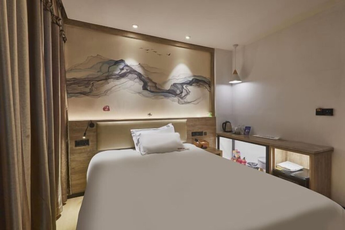 A hotel room with a large bed, a nightstand, a wall-mounted artwork, and a desk with a lamp.