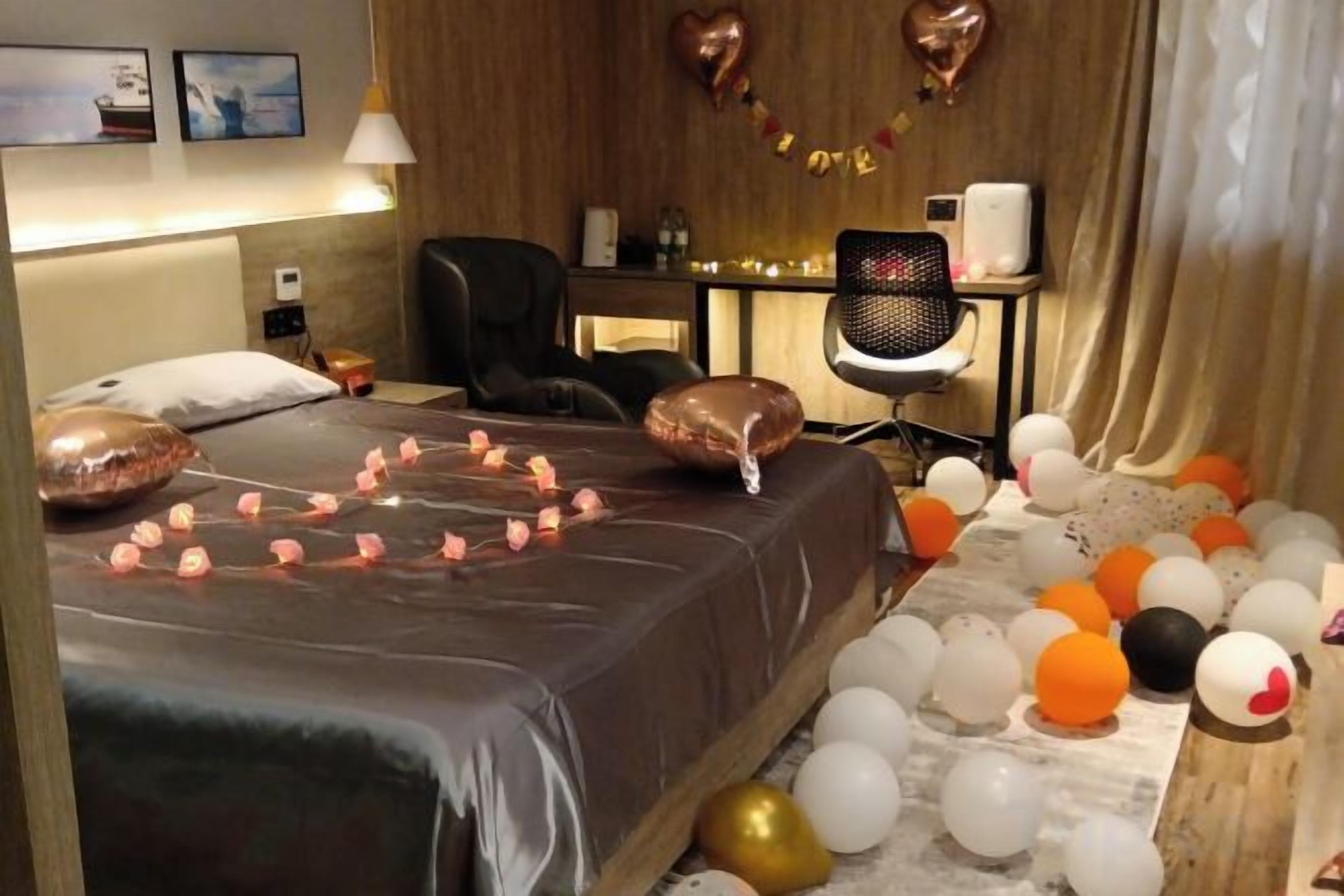 A hotel room with a bed, a desk, and a chair. There are balloons on the floor and a string of lights on the bed.