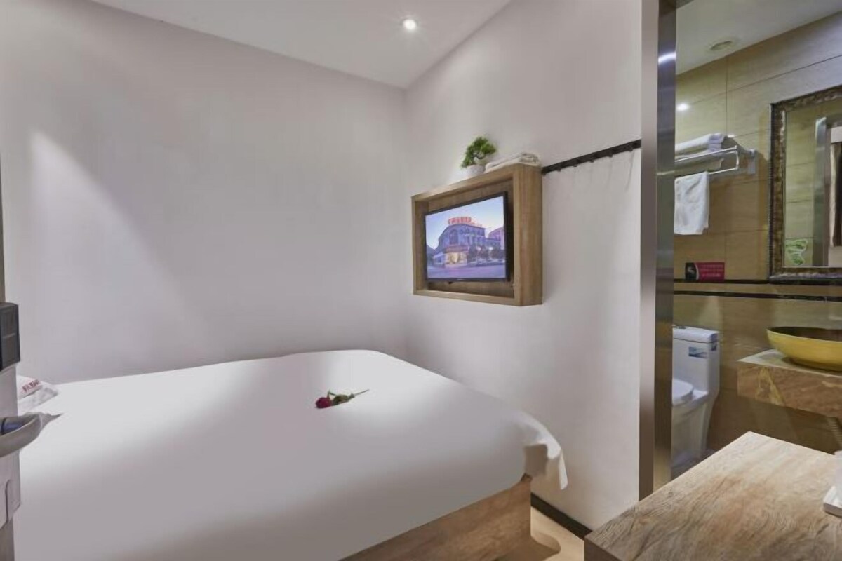A hotel room with a bed, a television mounted on the wall, a small plant, a bathroom with a mirror, and a sink.