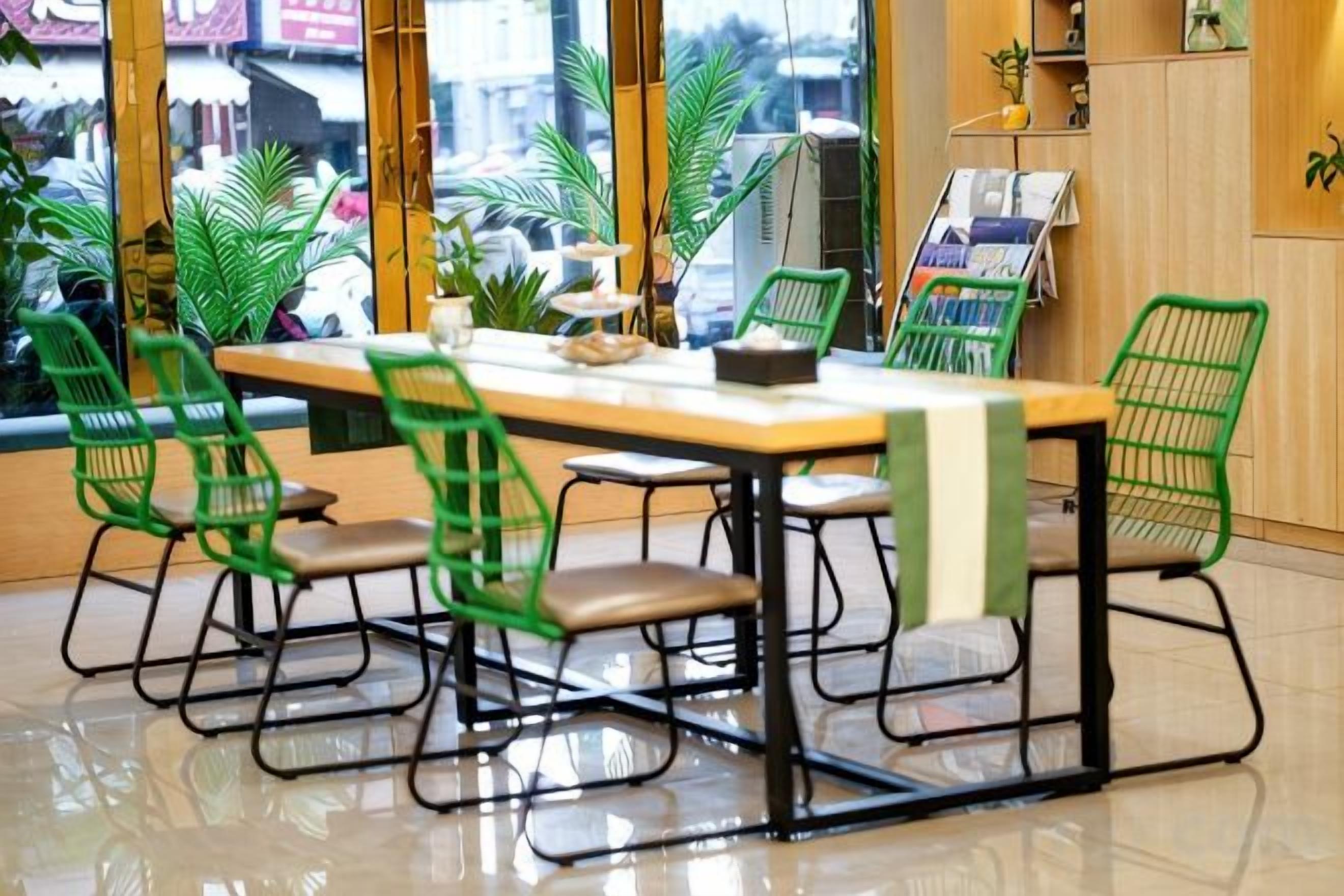 A dining area with a long table, green chairs, and a view through large windows.