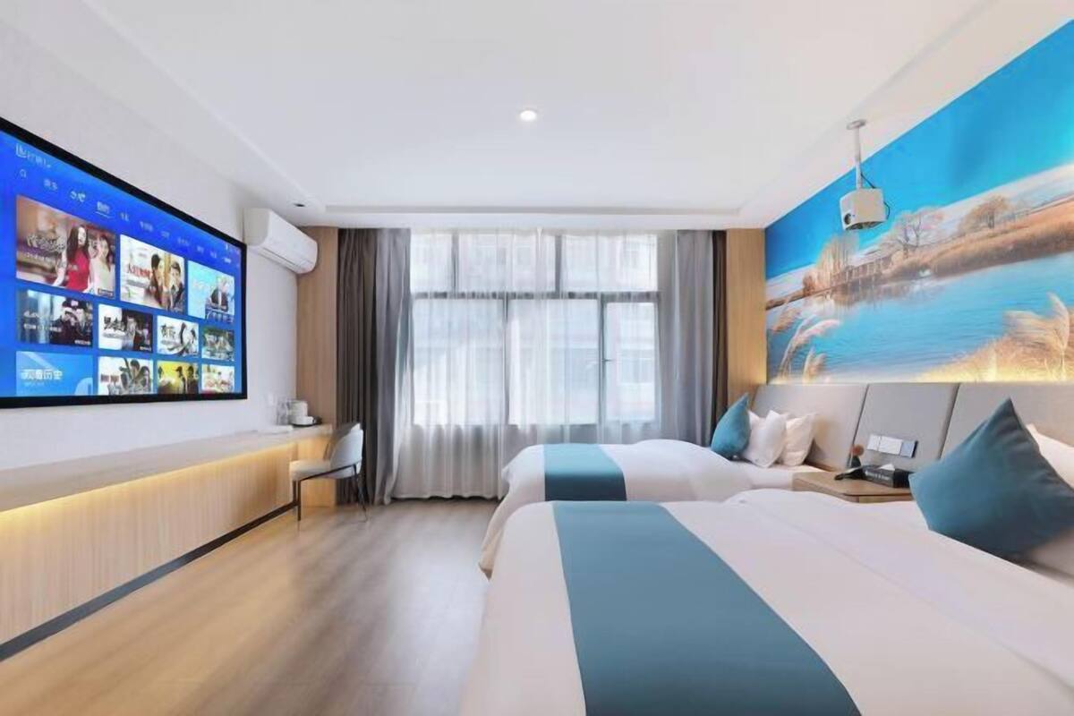 A hotel room with a large digital screen displaying content, a bed with a view of a painting, and a desk with a chair.
