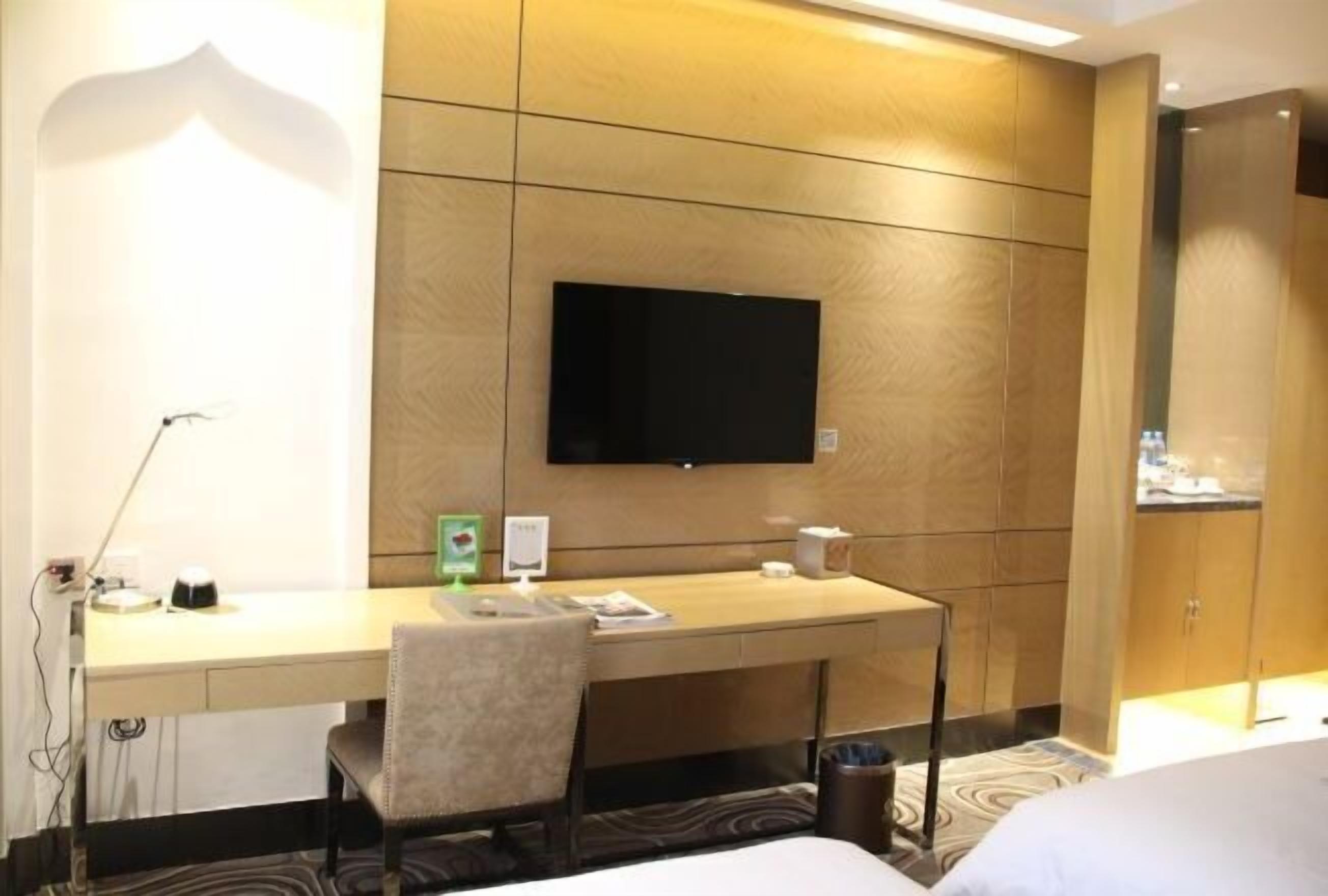 Una habitación de hotel con un escritorio de madera, una silla, un televisor de pantalla plana montado en la pared y una cama con ropa blanca.