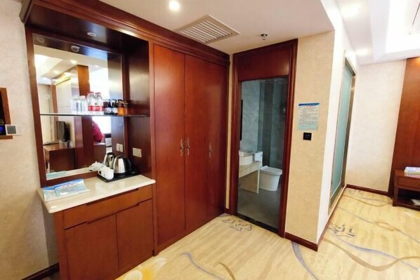 A hotel room with a wooden wardrobe, a mirror, a countertop with toiletries, and a bathroom visible through a glass door.