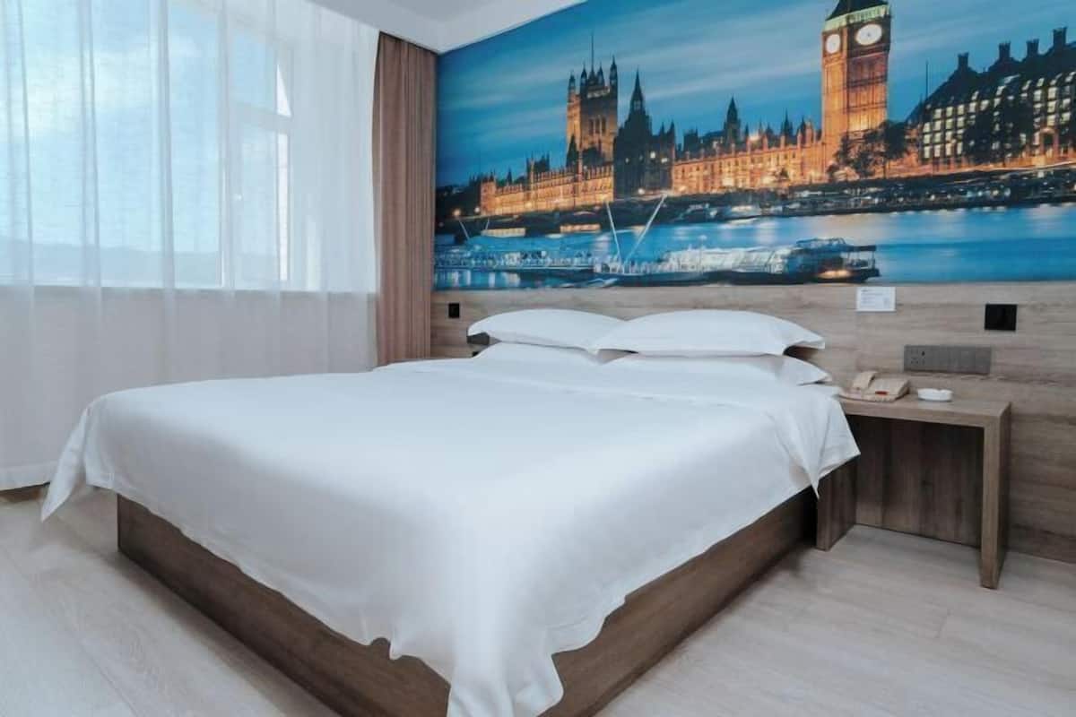 Ein Hotelzimmer mit einem großen Bett, einem Nachttisch, einem Stadtbild und einem Fenster mit Vorhängen.