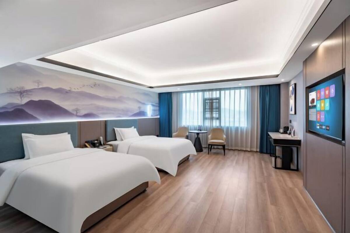 Quarto de hotel com duas camas, uma escrivaninha, uma cadeira, televisão na parede e um quadro grande.