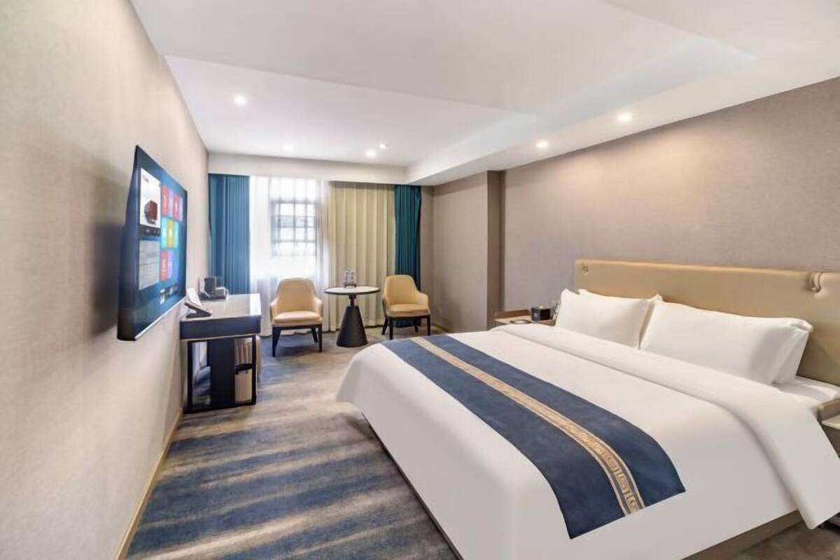 Quarto de hotel com cama, escrivaninha, duas cadeiras, televisão e janela com cortinas.