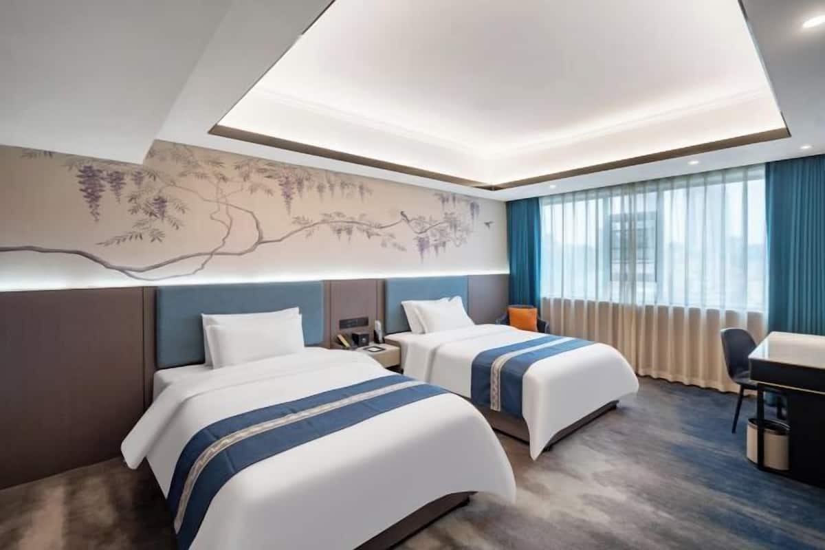 Quarto de hotel com duas camas, uma escrivaninha e uma janela grande com cortinas.