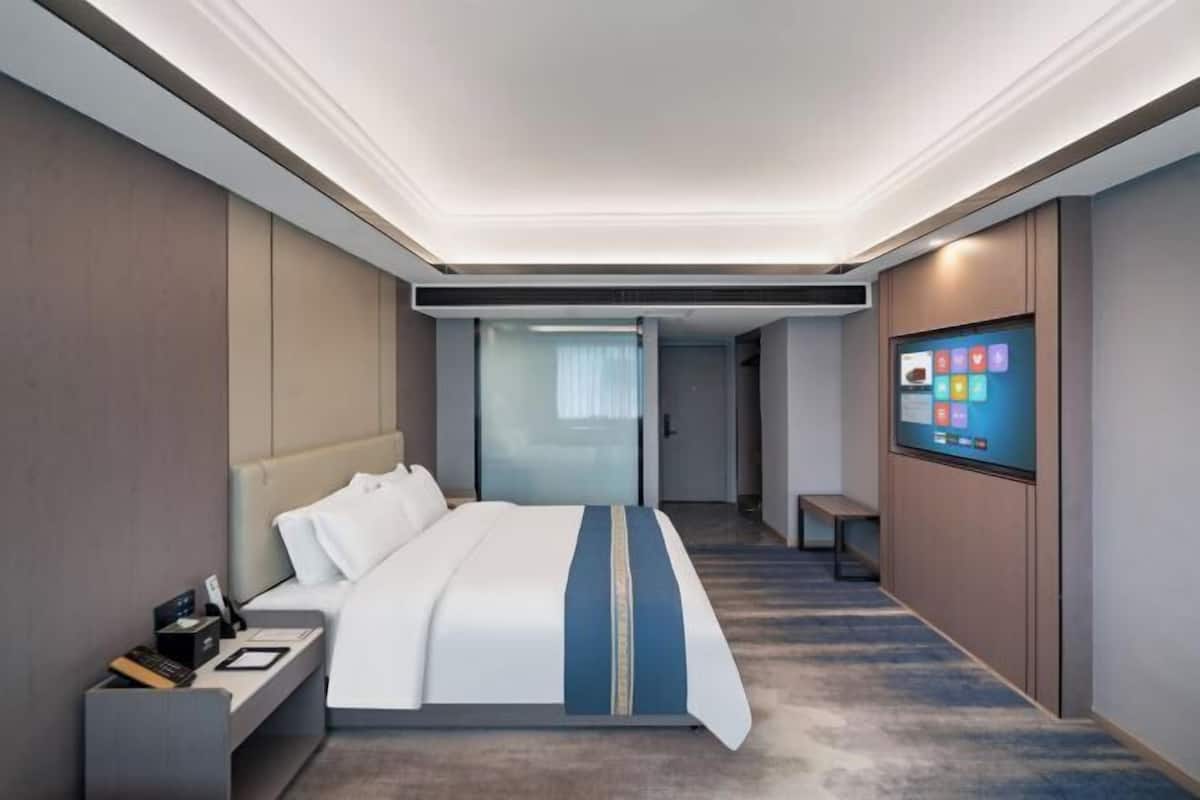 Quarto de hotel moderno com uma cama grande, uma escrivaninha com um computador, uma televisão na parede e um banco no canto.