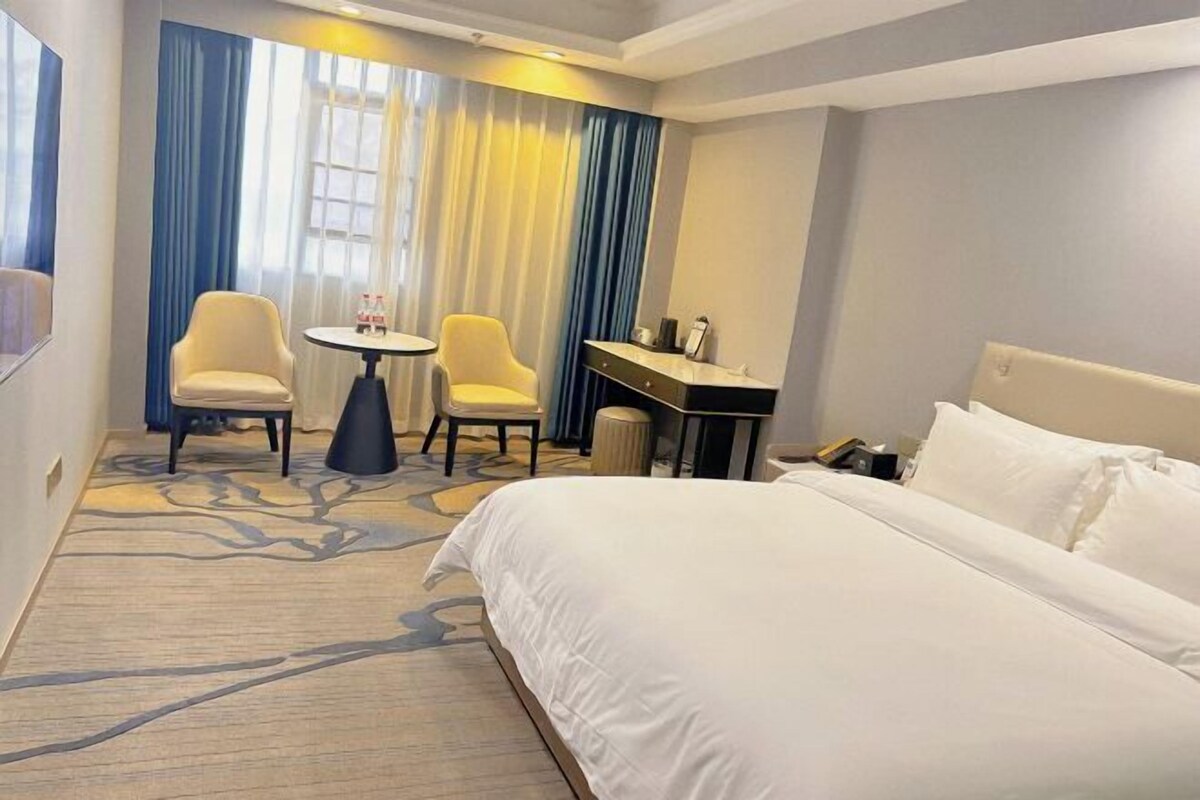 A hotel room with a large bed, two chairs, a small table, a desk, and a window with curtains.