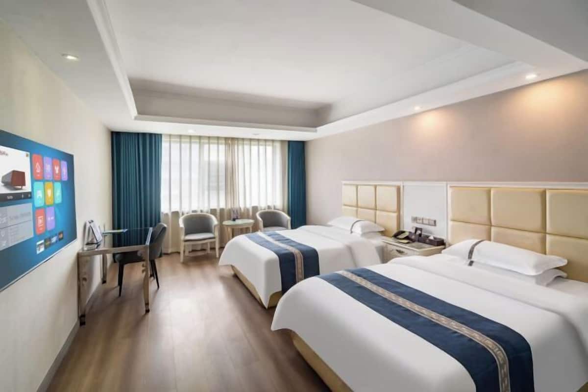 Quarto de hotel com duas camas, uma escrivaninha, uma cadeira, uma mesinha e uma televisão na parede.