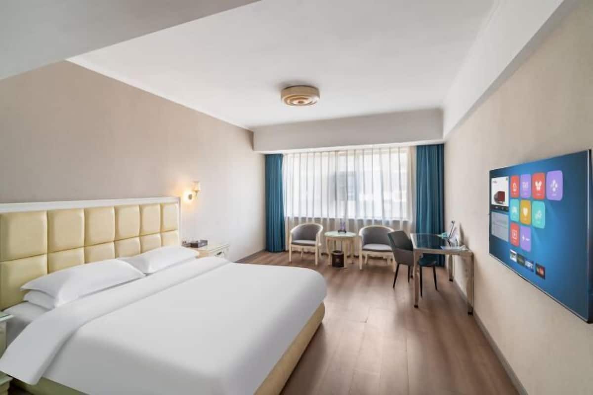 Quarto de hotel com uma cama grande, uma mesa, duas cadeiras, uma televisão e uma janela com cortinas.